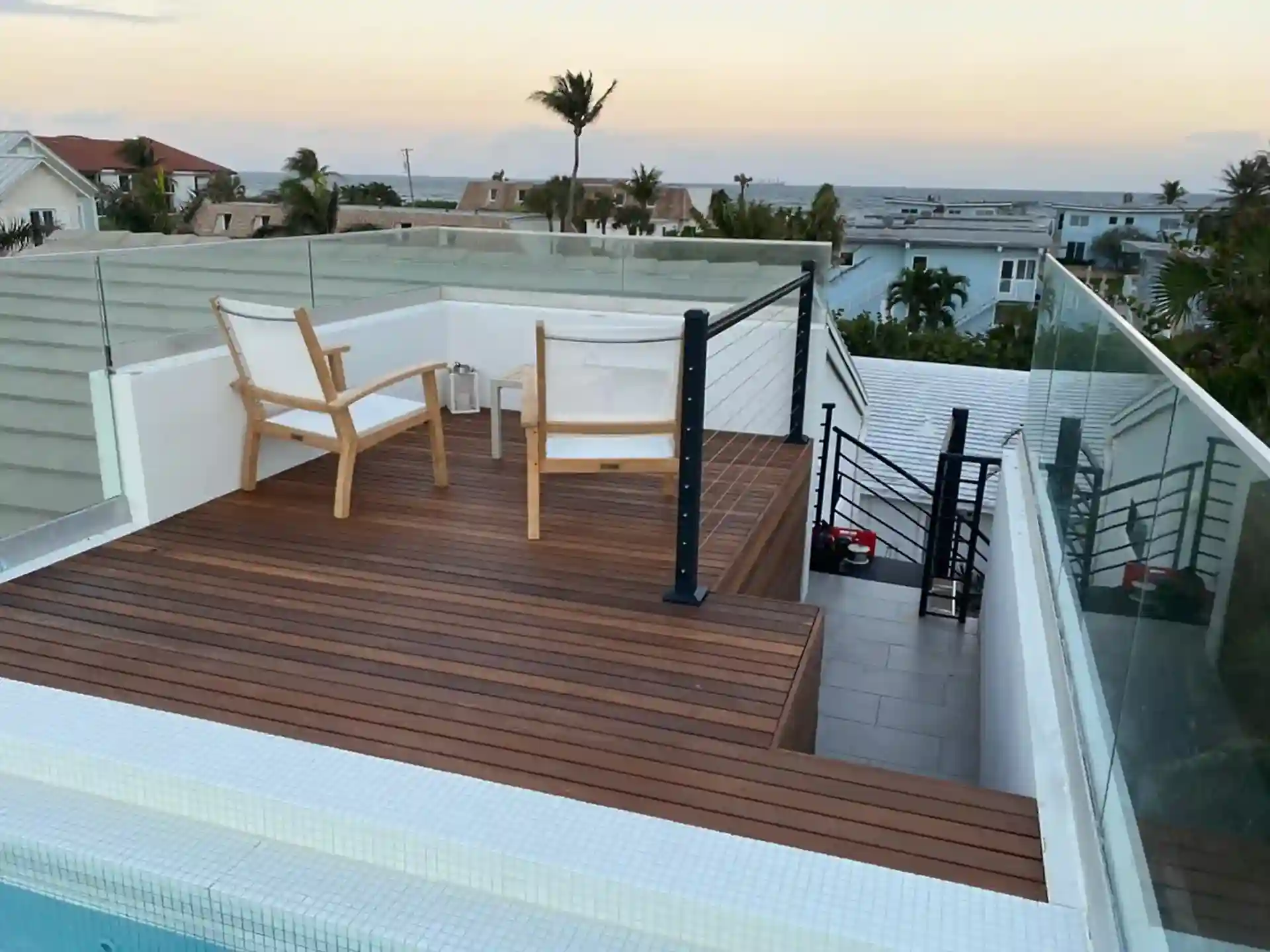 Photo of a rooftop deck with deck flooring, glass railings, and two chairs overlooking the ocean. 